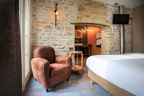 A hotel room with a stone wall, a bed, a chair, a small table with a water bottle, and a TV mounted on the wall. - Le Terminus (Tournus)