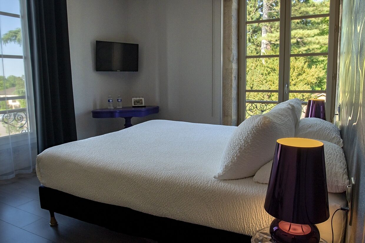 A neatly made bed with a white bedspread and pillows, a bedside table with a lamp, a flat-screen TV mounted on the wall, and a window with a view of greenery.