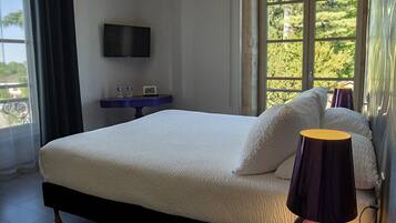 A neatly made bed with a white bedspread and pillows, a bedside table with a lamp, a flat-screen TV mounted on the wall, and a window with a view of greenery.