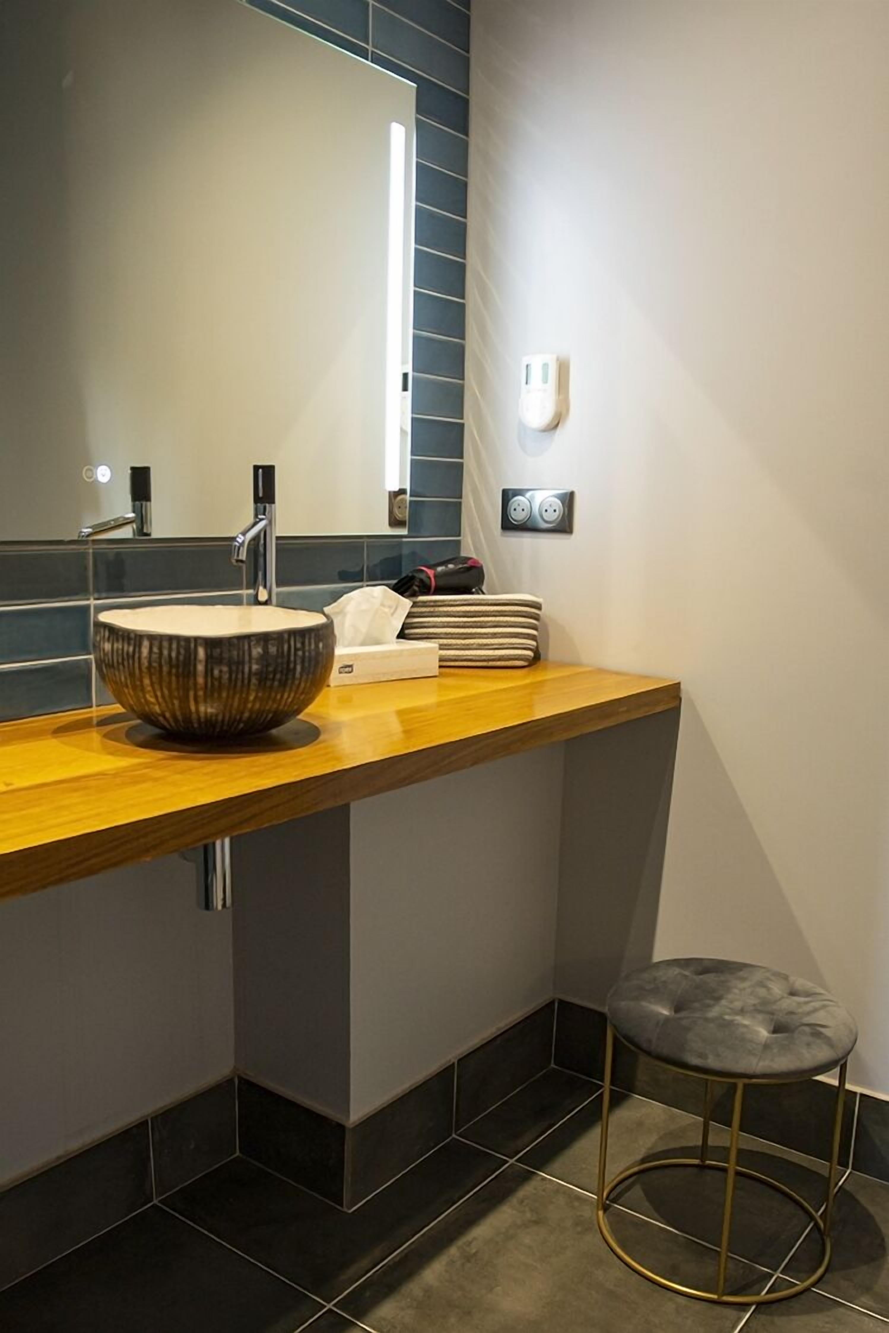 Une salle de bain moderne avec un grand lavabo sur pied, un plan de toilette en bois, un tabouret et un miroir doté d’une bande lumineuse.