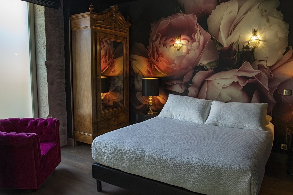 A bedroom with a large bed, a wooden wardrobe, a purple armchair, and a wall mural of flowers.