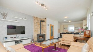 Una sala de estar moderna con una televisión de pantalla plana, una mesa de centro de vidrio, sofás color beige, una chimenea de piedra y una cocina al fondo.