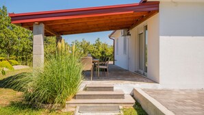 Un espacio exterior techado con una mesa y sillas, escalones que conducen a la entrada de un edificio, y vistas de la vegetación circundante.
