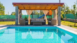 Una piscina con zona de estar techada y un tobogán, rodeada de vegetación.