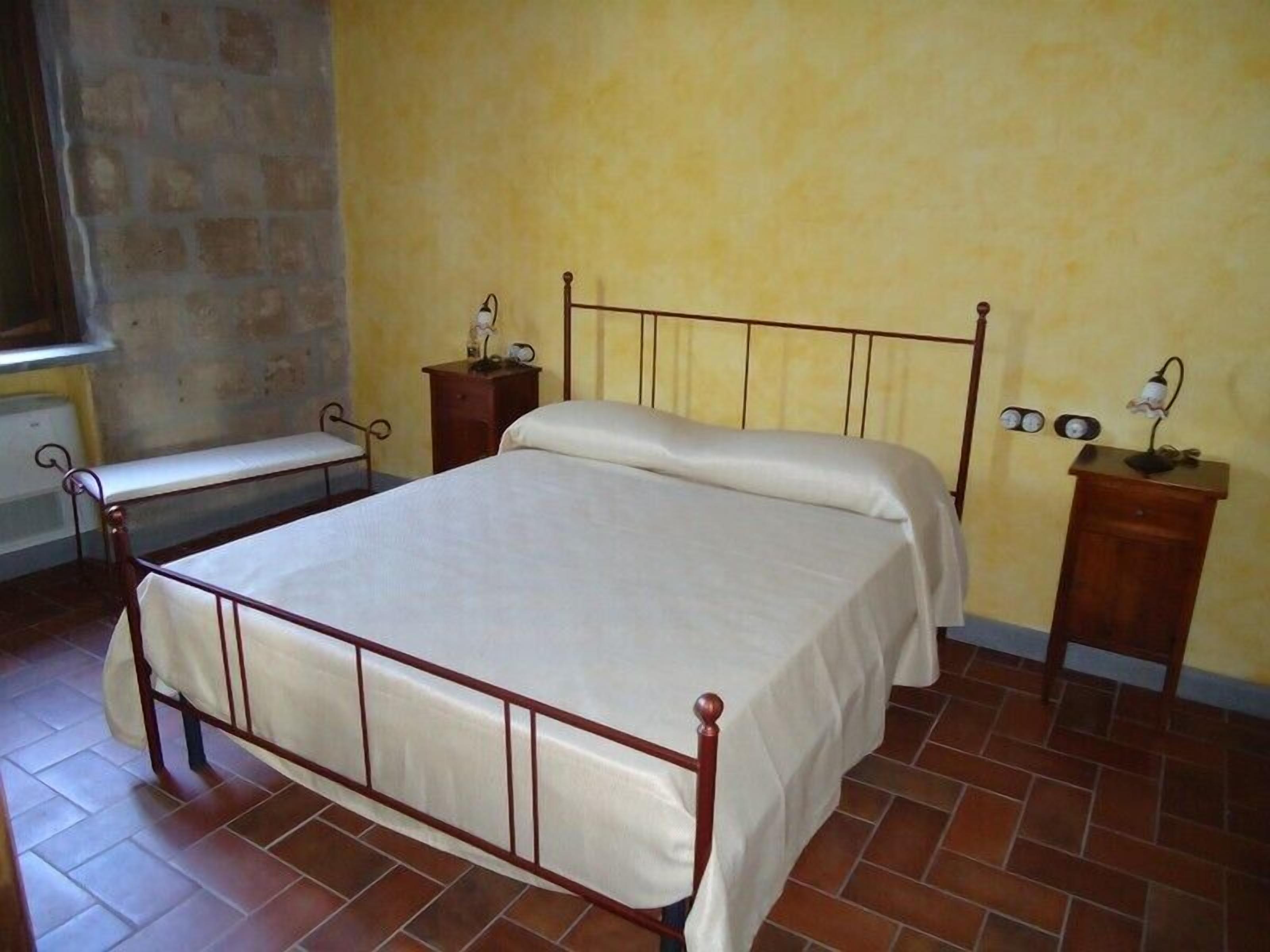 A bedroom with a metal-framed bed, a bedside table with a lamp, and a bench against the wall.