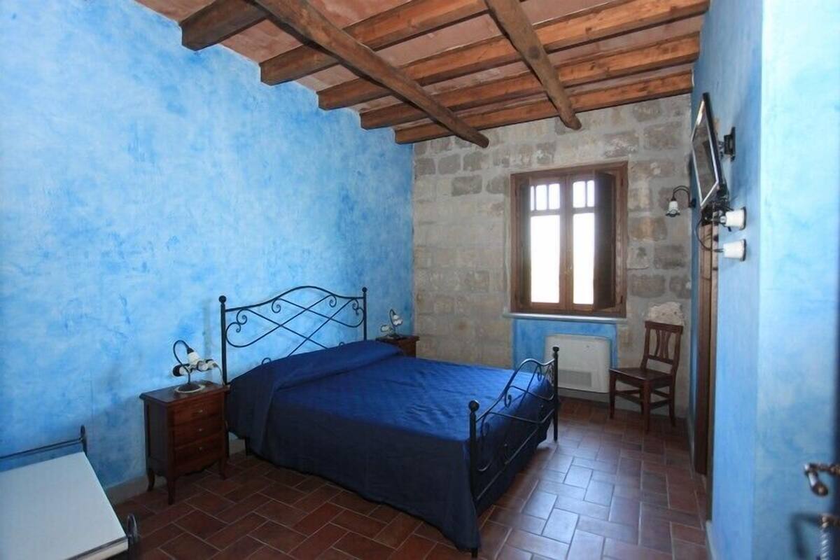 Un dormitorio con cama, techo de madera, pared de piedra, ventana y una silla.