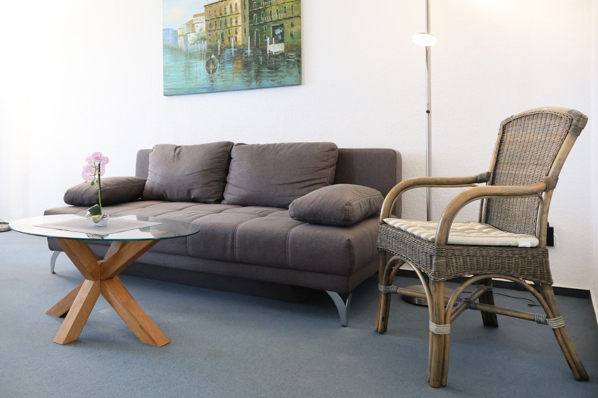 A modern living room with a grey sofa, a wicker chair, a glass coffee table, and a painting of a canal scene on the wall.