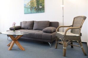 En moderne stue med en grå sofa, en fletstol, et glasbord og et maleri af et kanalmotiv på væggen.