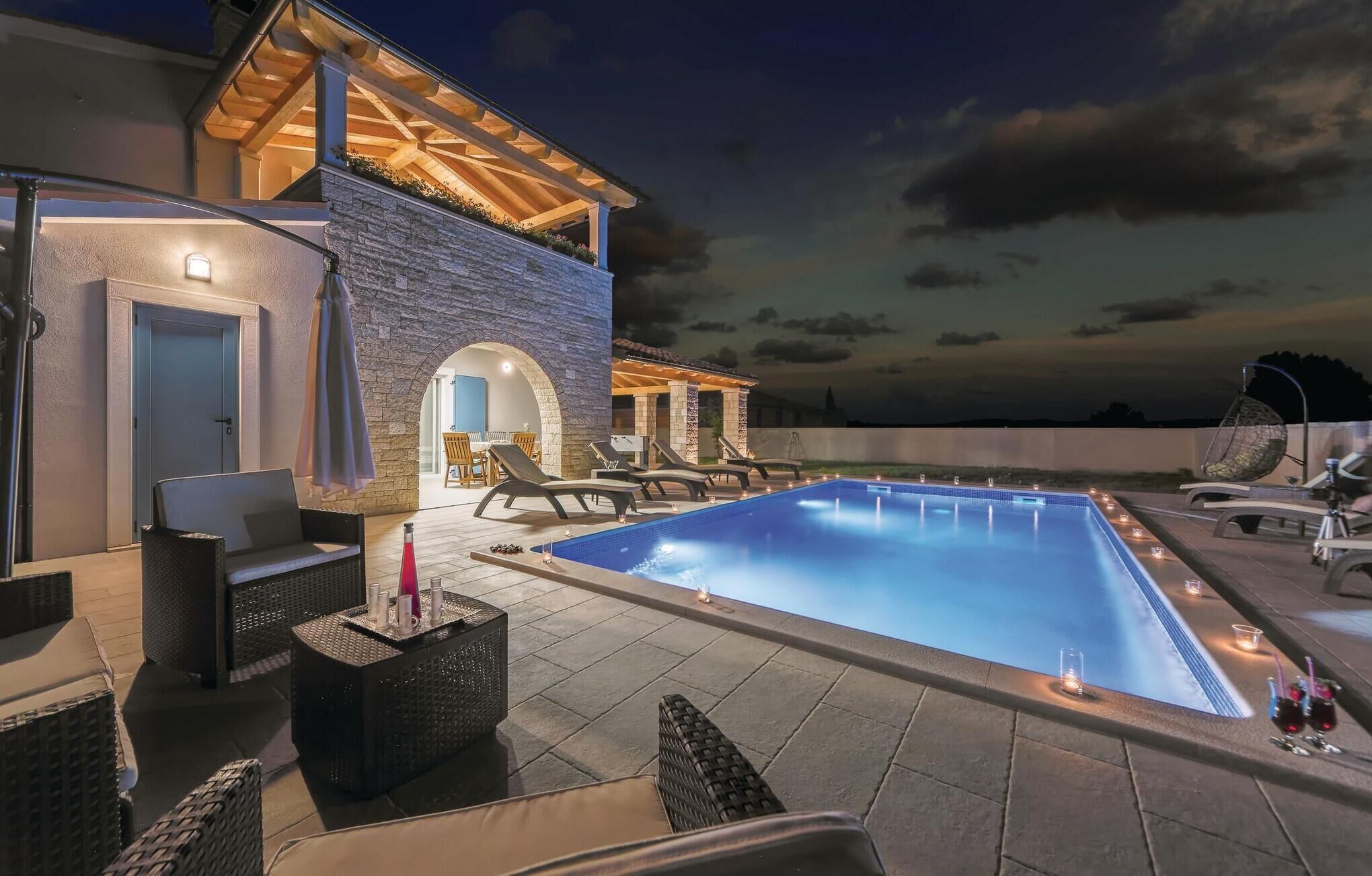Une piscine dotée de chaises longues et d’un espace salon couvert, le tout entouré de murs en pierre et d’une pergola en bois.