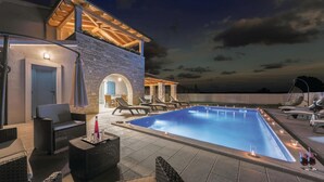 Une piscine dotée de chaises longues et d’un espace salon couvert, le tout entouré de murs en pierre et d’une pergola en bois.