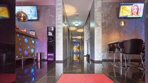 Un hall d’hôtel avec un tapis rouge sur lequel on peut lire « HÔTEL PLATIN », un comptoir d’accueil, un écran de télévision et un espace bar.