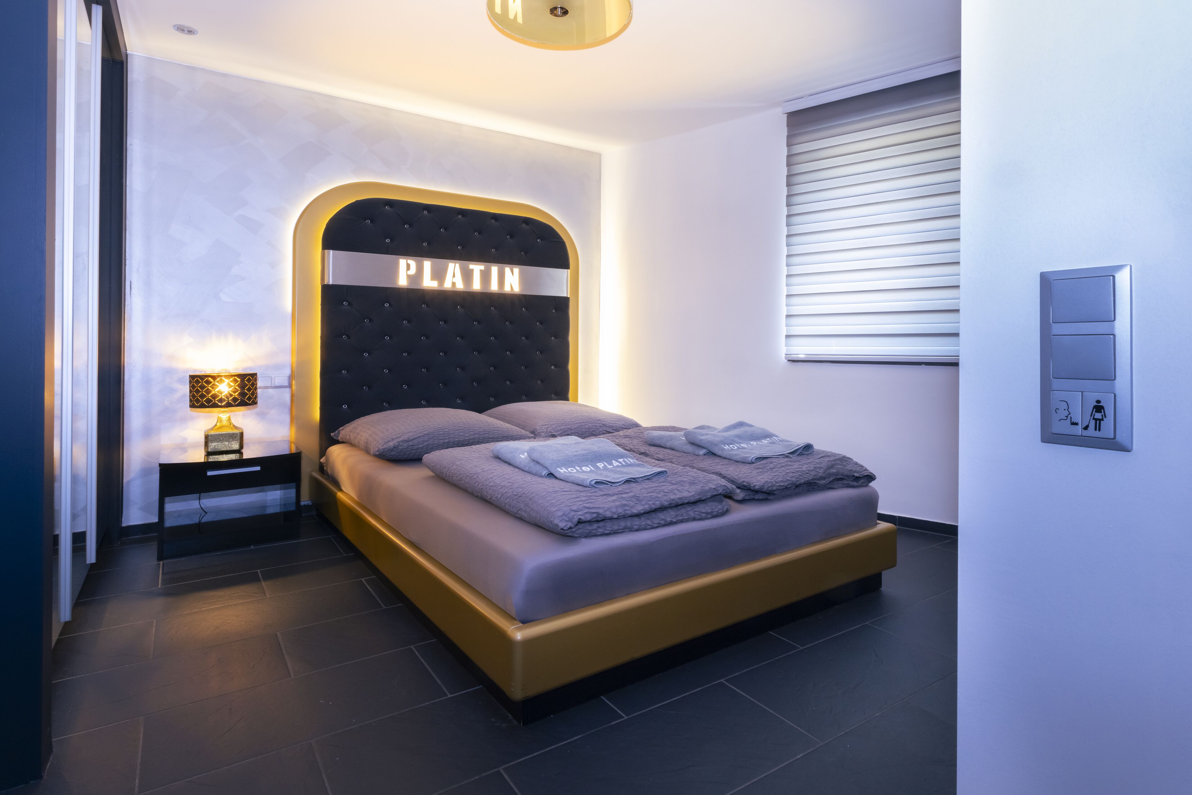 A modern hotel room with a large bed, a headboard with 'PLATIN' branding, a bedside table with a lamp, and a window with blinds.
