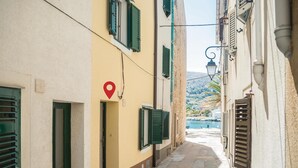 A narrow street with buildings on either side, a red location marker, and a view of the sea with boats in the distance.