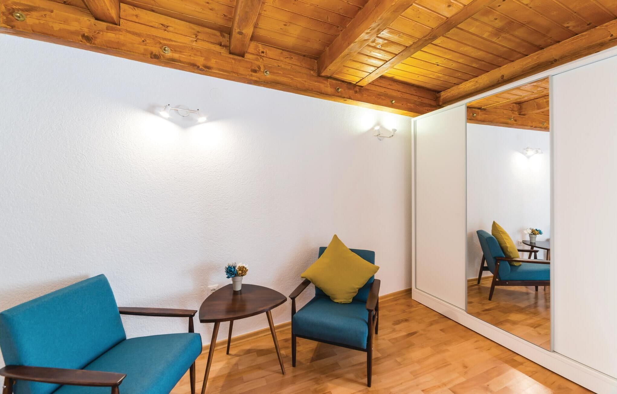 A room with wooden flooring, a blue sofa, a small table with a vase, and a mirror reflecting a chair and a table with yellow cushions.