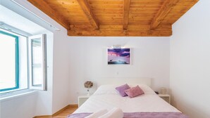 A bedroom with a wooden ceiling, a bed with white and purple bedding, a nightstand, a vase with flowers, and a painting of a landscape.