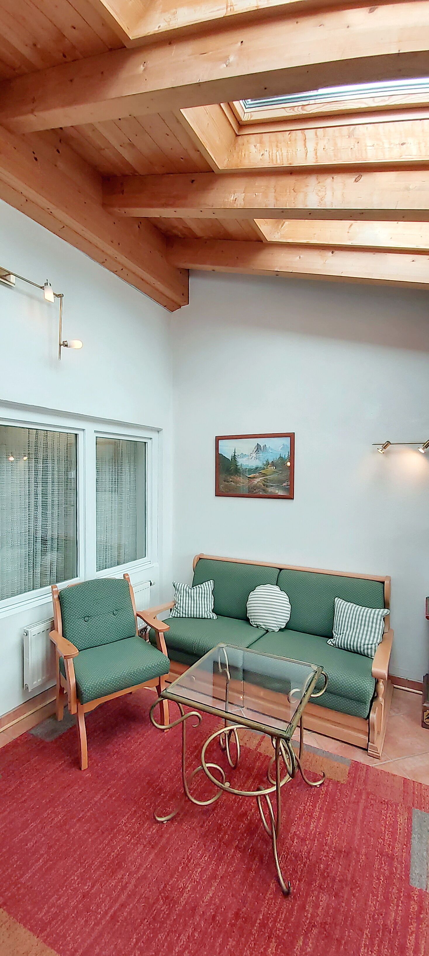 A cozy room with a wooden ceiling, a green sofa, a wooden chair, a glass table, and a red carpet.