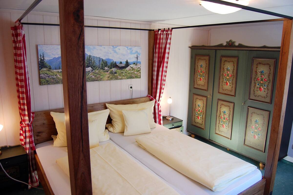 Une chambre à coucher avec une tête de lit en bois, un lit recouvert de draps blancs, une table de chevet avec une lampe, un tableau représentant un paysage accroché au mur, et une armoire verte ornée de panneaux décoratifs.