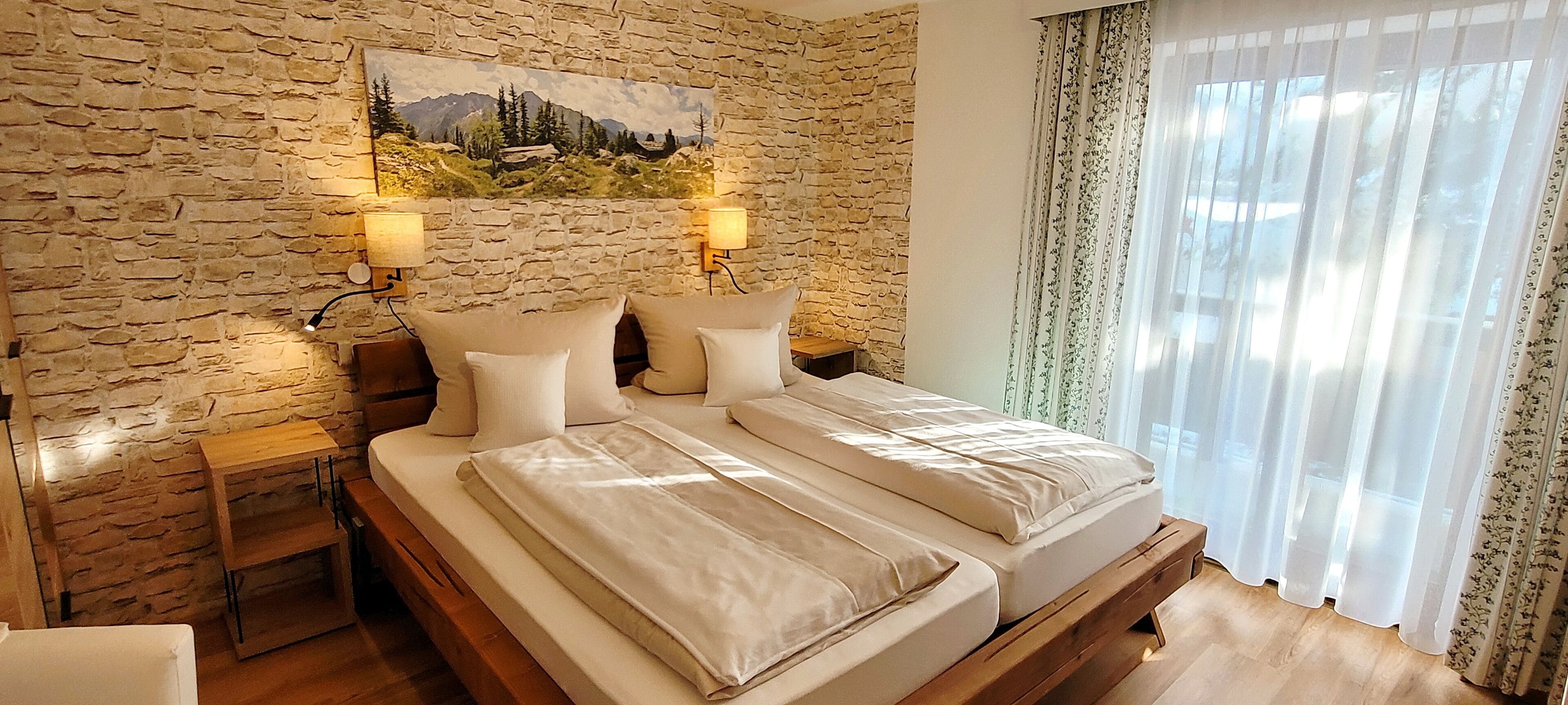 Una cama doble con cabecera de madera y ropa de cama color beige, una pared de piedra con un cuadro, dos lámparas de pared y una ventana con cortinas de encaje.
