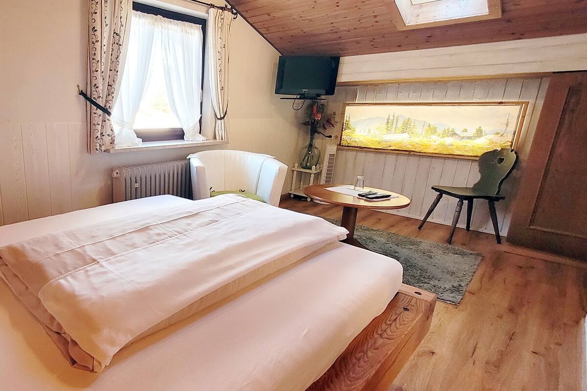 Une chambre à coucher avec un lit, une chaise, une petite table, un téléviseur et une fenêtre avec des rideaux.