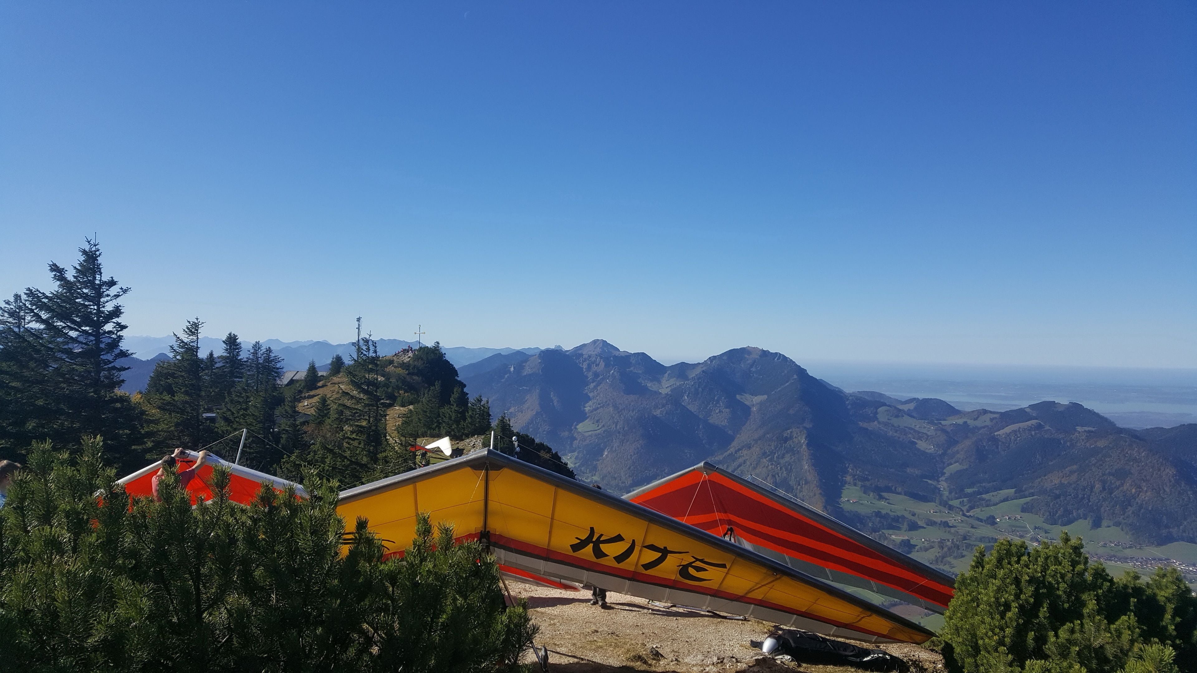 Eine Berglandschaft mit blauem Himmel. Zwei kleine Gebäude mit roten und gelben Dächern sind von Bäumen umgeben.