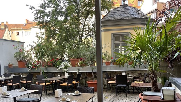 Un patio al aire libre con varias mesas y sillas, rodeado de plantas en macetas y un muro de piedra.
