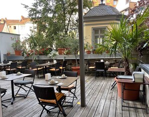 En udendørs terrasse med flere borde og stole, omgivet af plantekasser og en stenvæg.