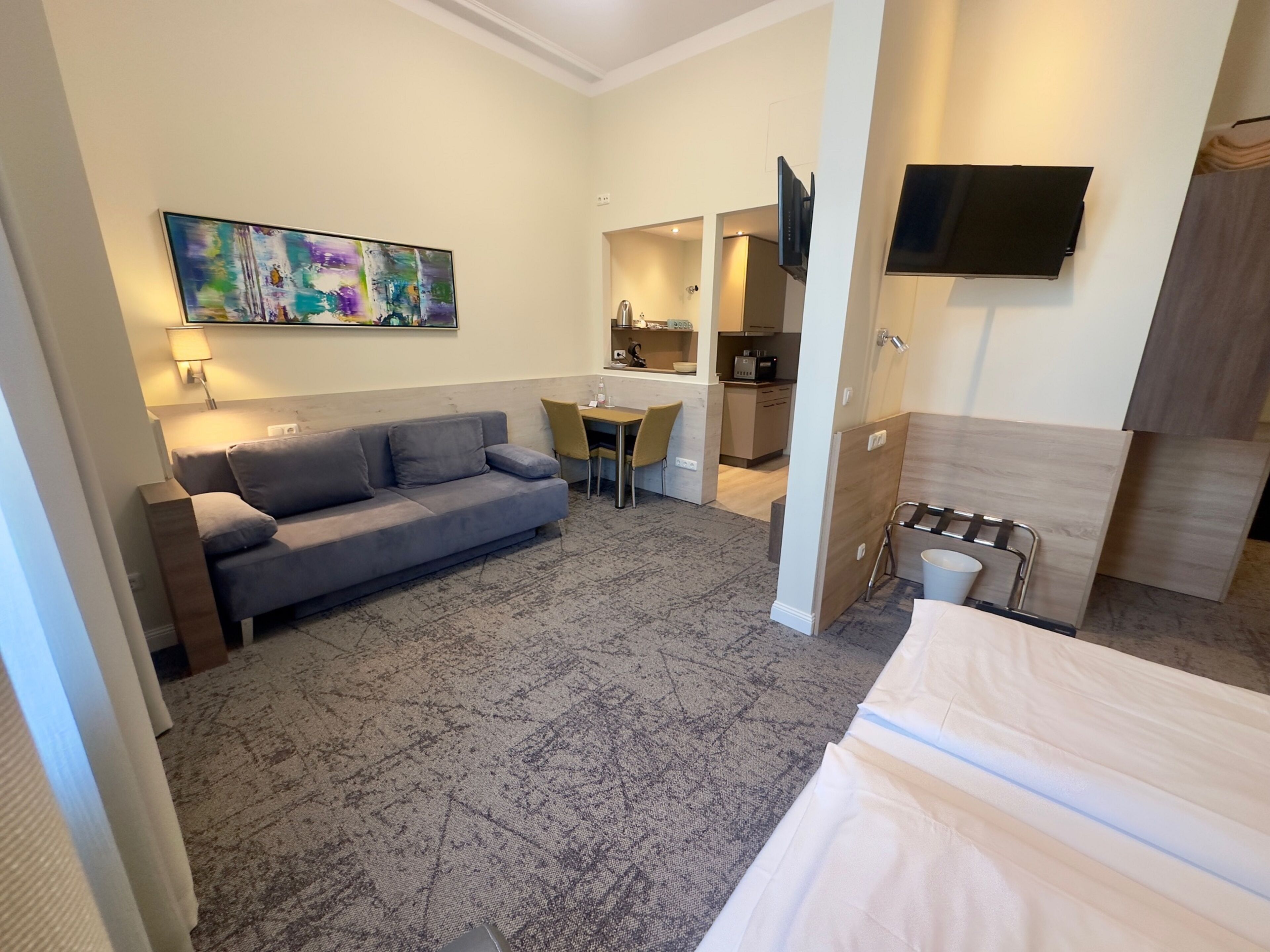 A compact hotel room with a grey sofa, a small table, a TV mounted on the wall, and a kitchenette in the background.