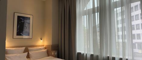 A hotel room with a bed, a wall-mounted light, and a window with sheer curtains.