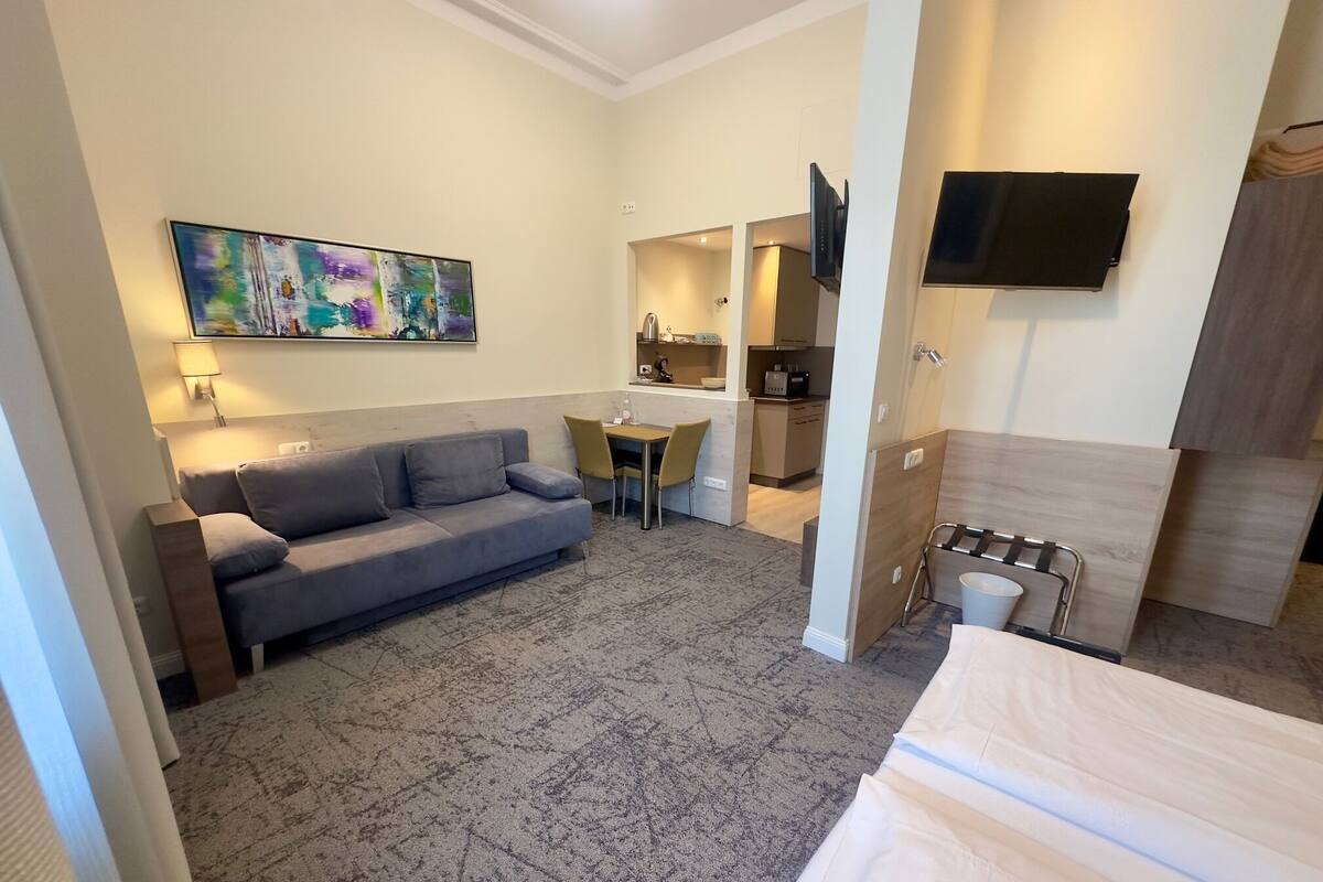 Una habitación de hotel moderna con un sofá gris, una pequeña mesa de comedor, un televisor montado en la pared y una zona de kitchenette.