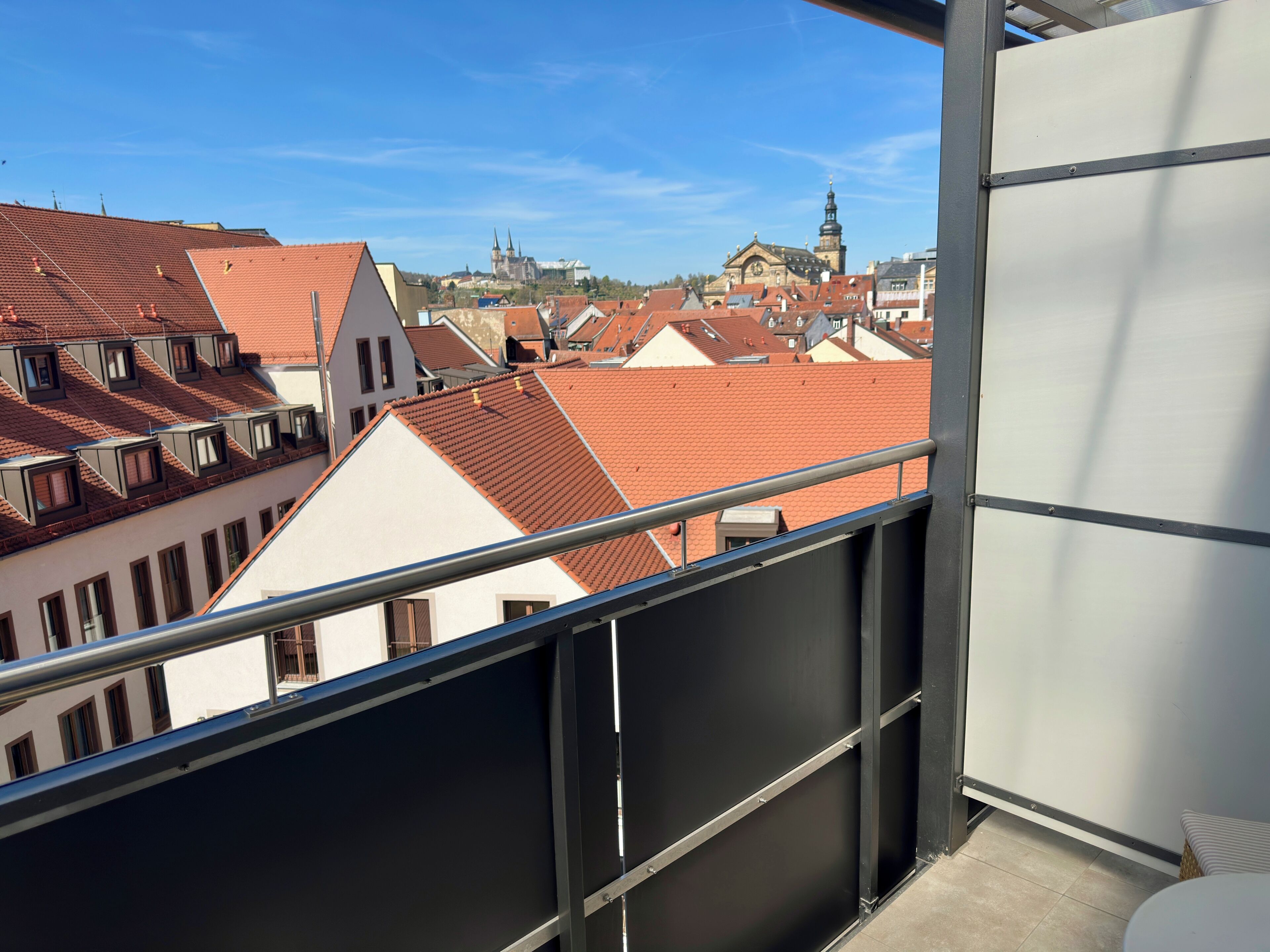 Ein Balkon mit Blick auf roten Ziegeldächern und eine Kirche mit zwei Türmen im Hintergrund.