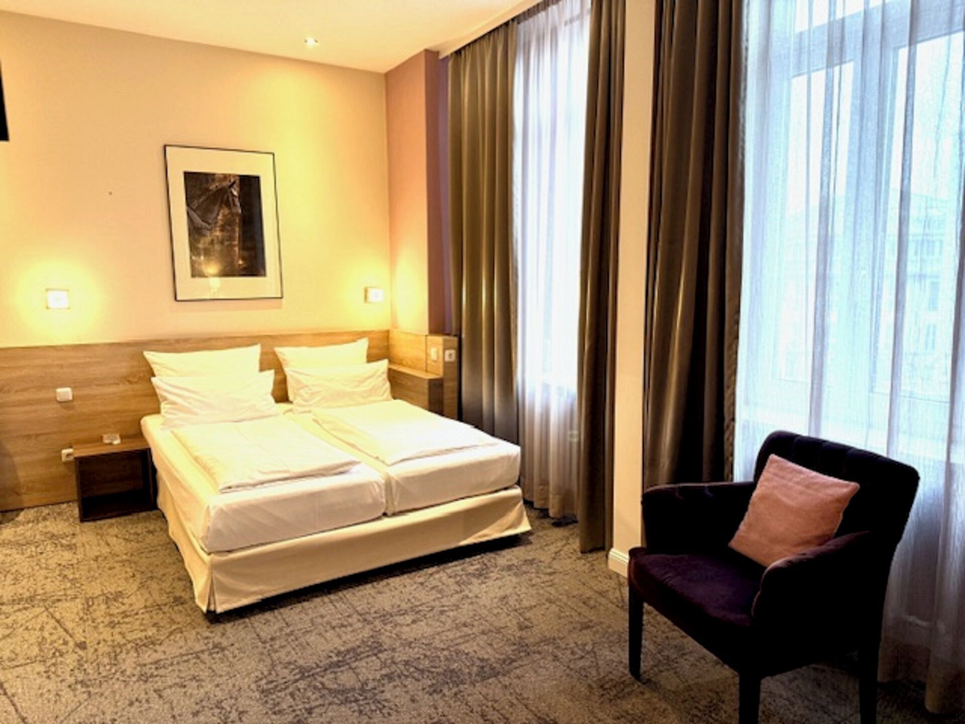 Ein Hotelzimmer mit Doppelbett, zwei Nachttischen, einem Sessel und einem großen Fenster mit Vorhängen. An der Wand hängt ein gerahmtes Bild.