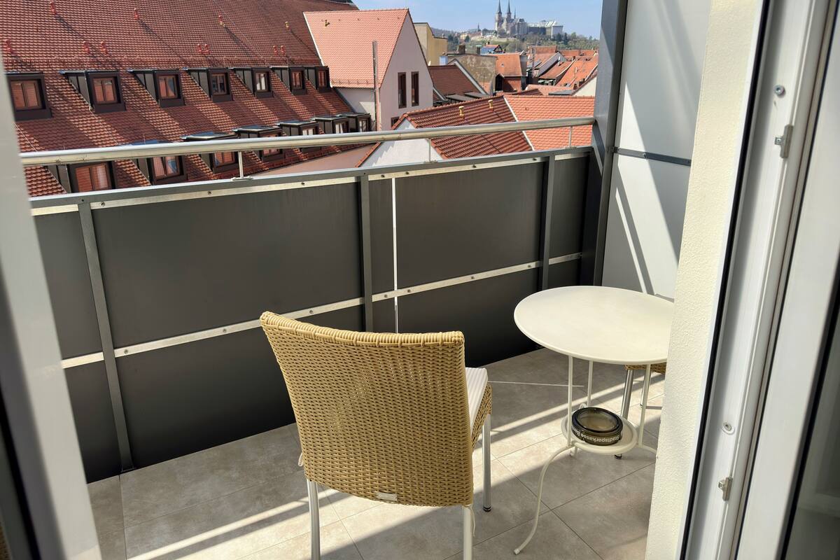 Un balcón con una silla de mimbre y una mesa pequeña, con vista a un paisaje urbano con edificios de techo rojo y una iglesia a lo lejos.