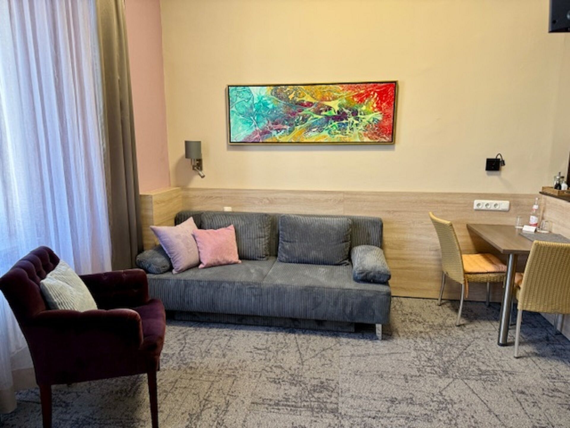 A hotel room with a gray sofa, a red armchair, a dining table with chairs, a colorful abstract painting on the wall, and a window with curtains.