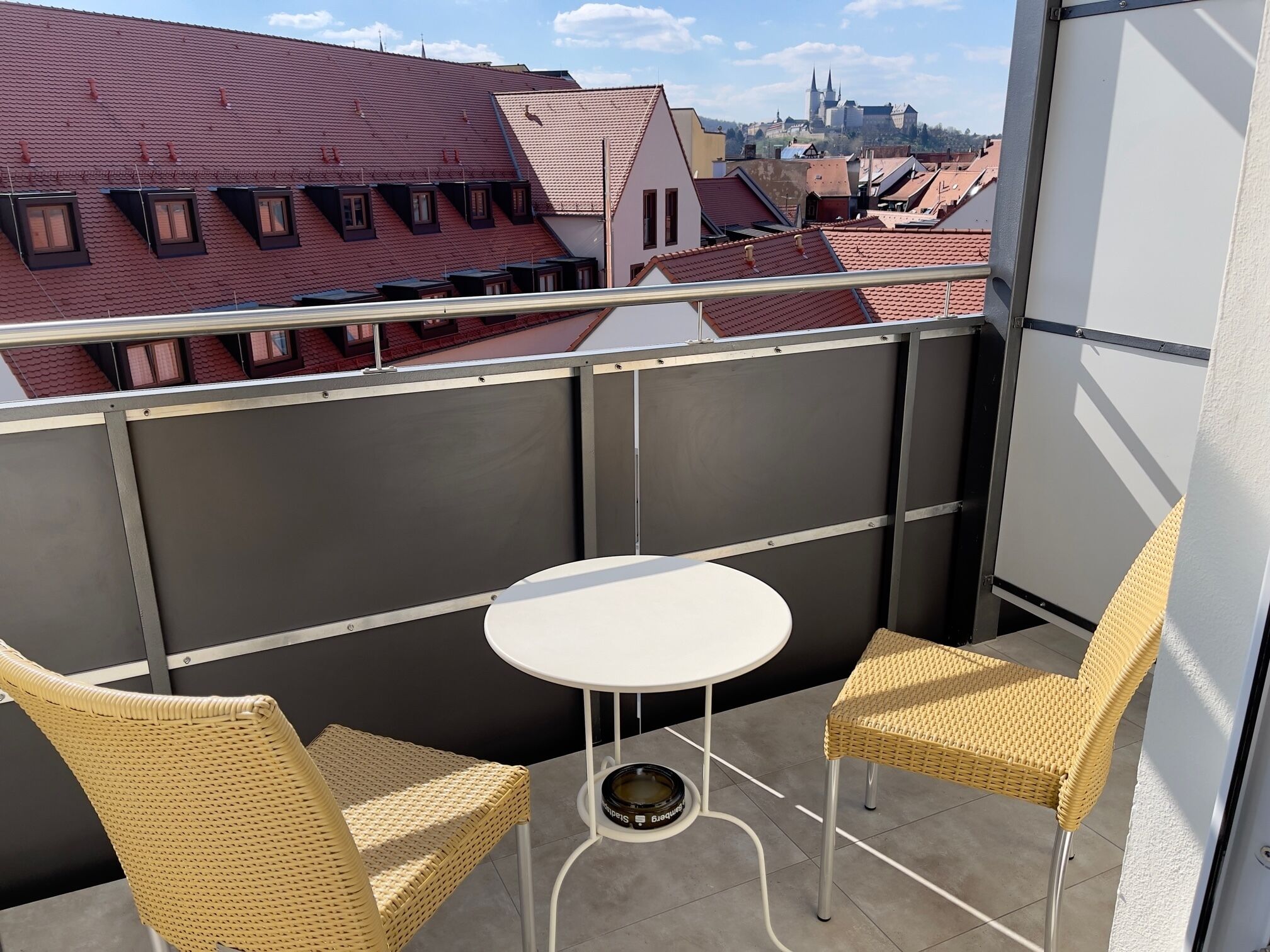 Ein Balkon mit zwei Korbstühlen und einem kleinen weißen Tisch, mit Blick auf eine Stadtlandschaft mit roten Dächern und einer in der Ferne sichtbaren Burg.