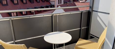 Ein Balkon mit zwei Korbstühlen und einem kleinen weißen Tisch, mit Blick auf eine Stadtlandschaft mit roten Dächern und einer in der Ferne sichtbaren Burg.