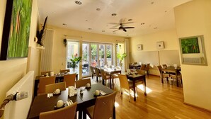 Comedor con varias mesas y sillas, ventilador de techo y vistas al exterior a través de amplios ventanales.