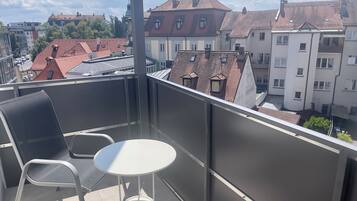 Un balcon avec une table et des chaises, donnant sur un paysage urbain composé de différents bâtiments et d’un ciel dégagé.