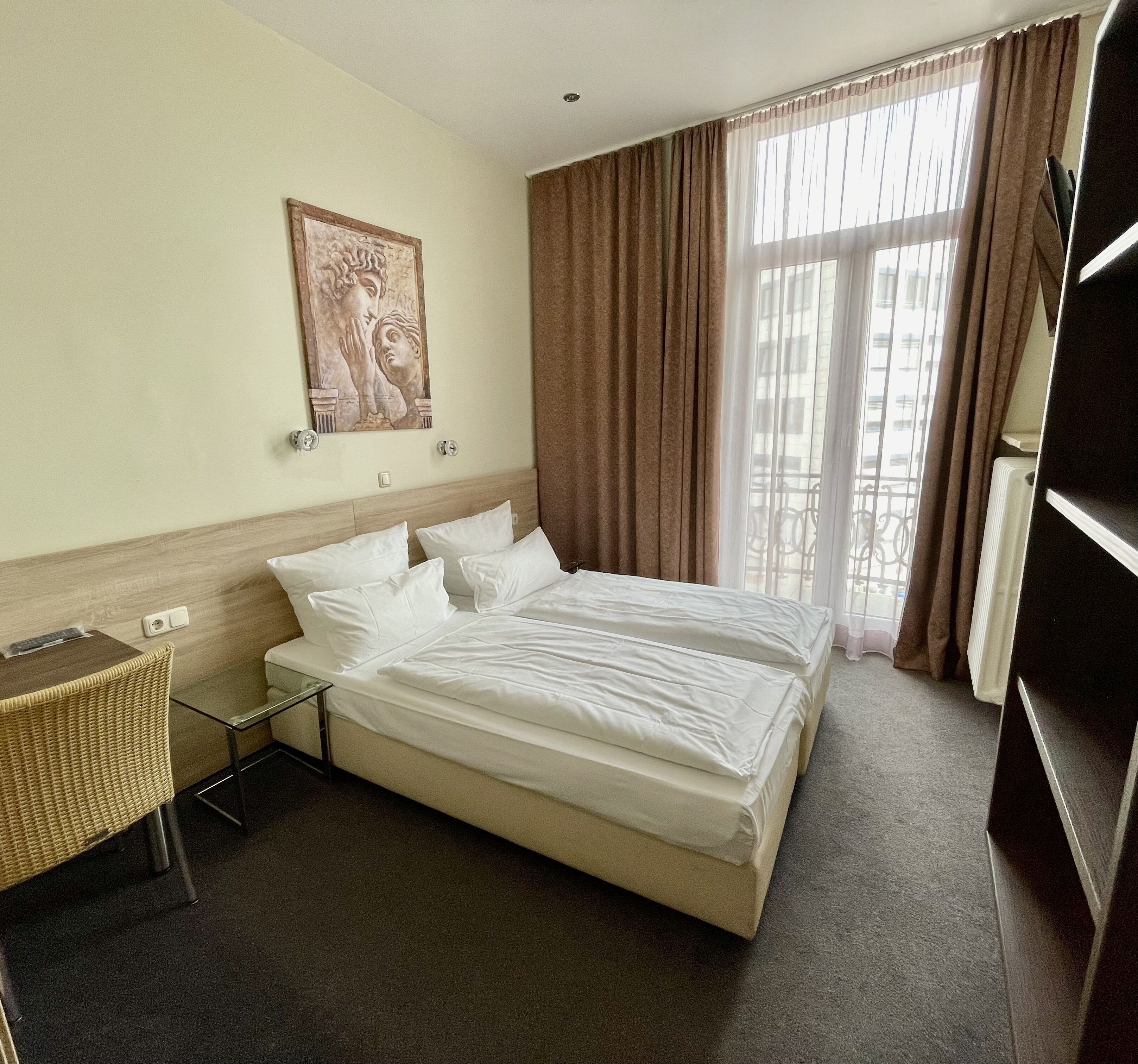 A hotel room with a bed, a chair, a table, a painting, and a window with curtains.