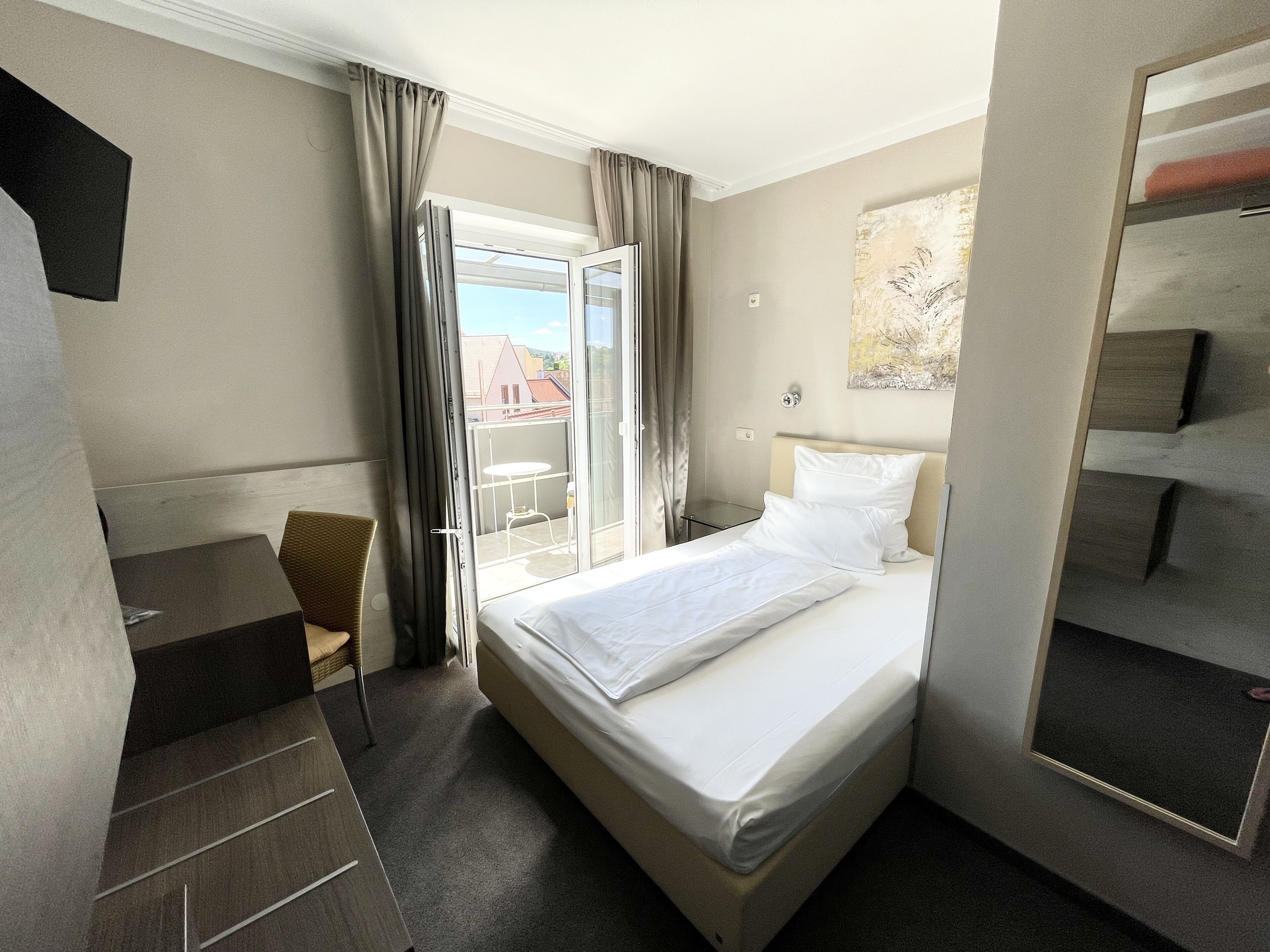 A hotel room with a bed, a desk, a chair, a large window with curtains, and a painting on the wall.