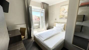 A hotel room with a bed, a desk, a chair, a large window with curtains, and a painting on the wall.