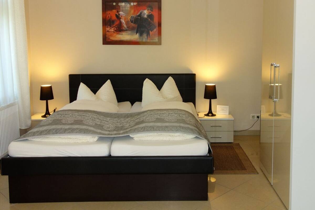 A neatly made bed with a black headboard, two bedside tables with lamps, a framed artwork above the bed, and a red chair to the left.