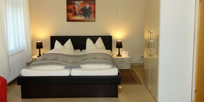 Un lit bien fait, avec une tête de lit noire, deux tables de chevet avec des lampes, une œuvre d’art encadrée au-dessus du lit et une chaise rouge sur le côté gauche.