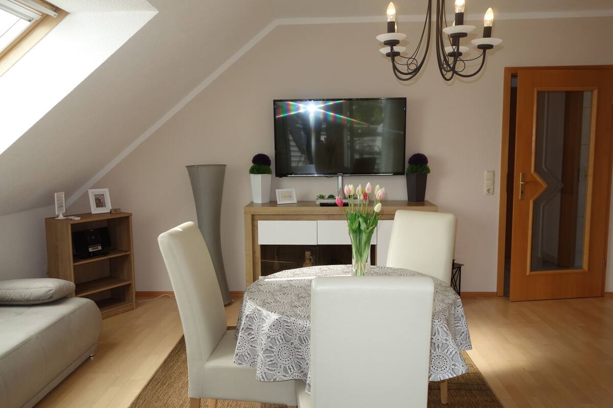 A cozy room with a TV mounted on the wall, a dining table with a lace tablecloth, a sofa, and a wooden cabinet.