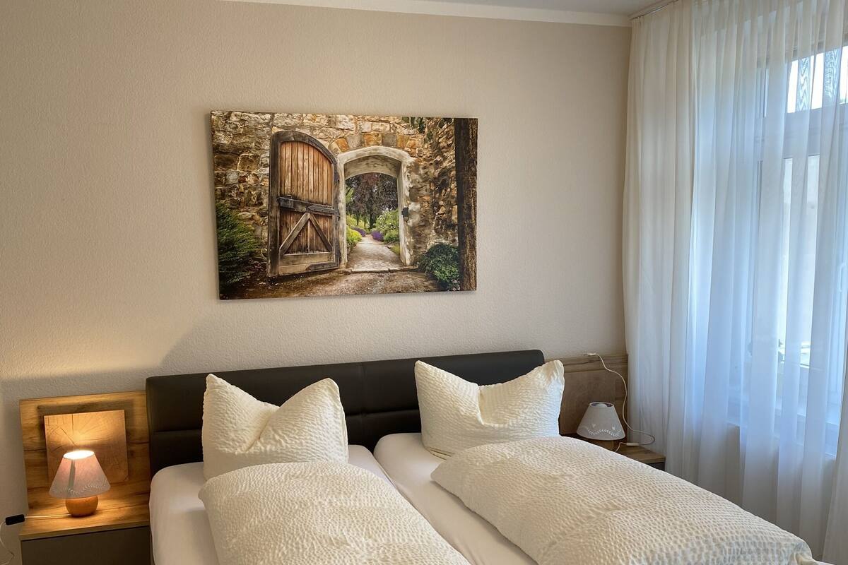 A double bed with white bedding, a nightstand with a lamp, a painting of a stone archway, and a window with sheer curtains.
