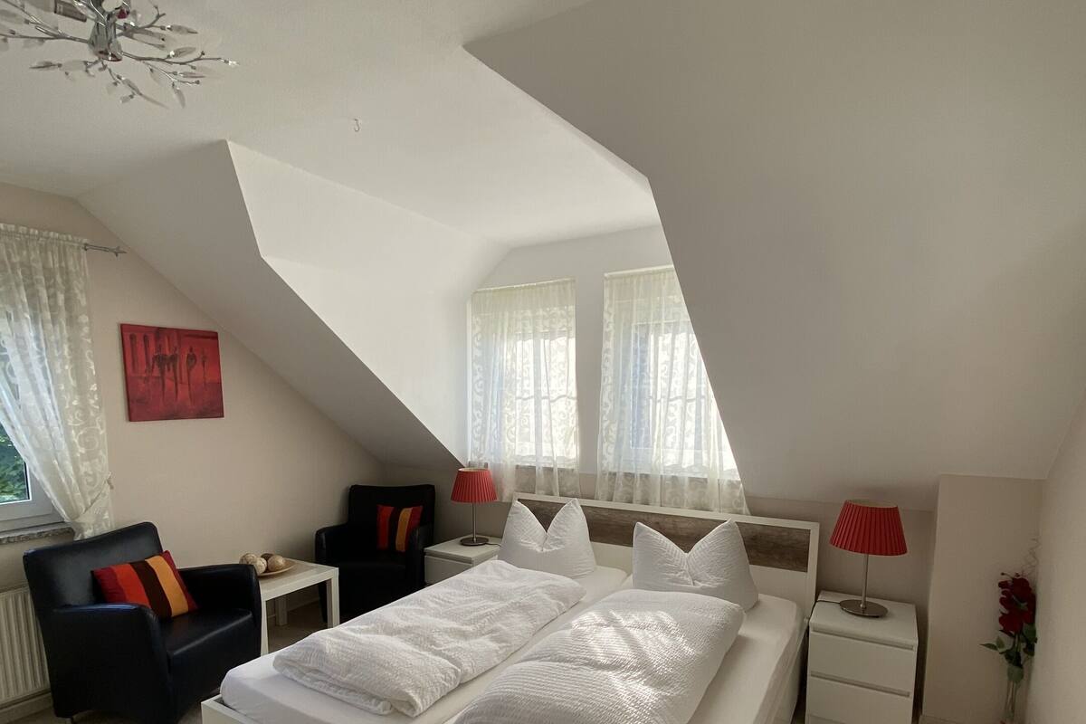 A modern bedroom with a large bed, a small table, a chair, and a nightstand.