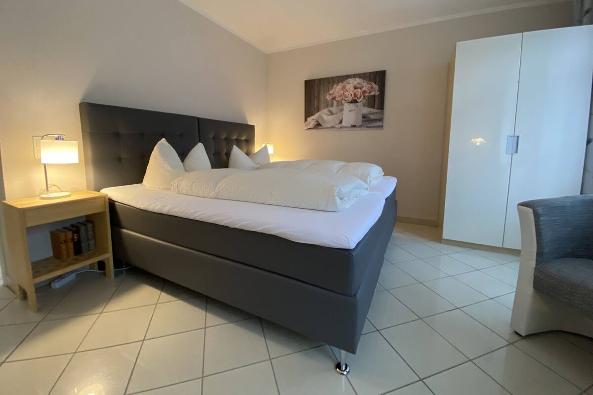 A modern bedroom with a large bed, a nightstand, a chair, and a wardrobe.