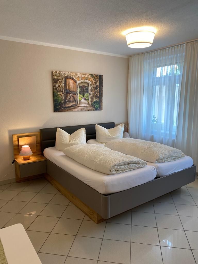 A bedroom with a bed, a nightstand with a lamp, a painting on the wall, and a window with curtains.