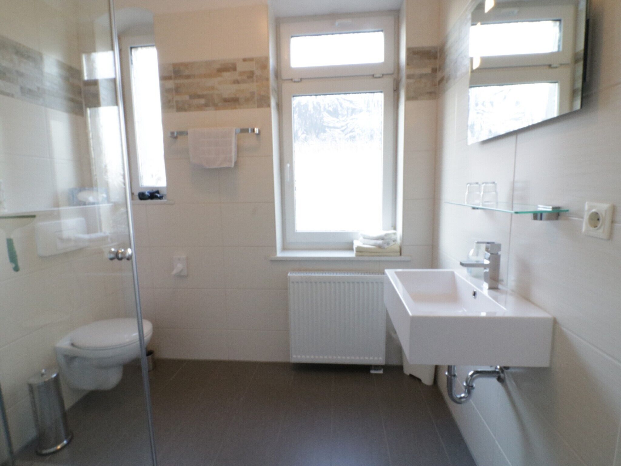 A modern bathroom with a glass shower enclosure, a white toilet, a large window, and a white sink with a mirror above it.