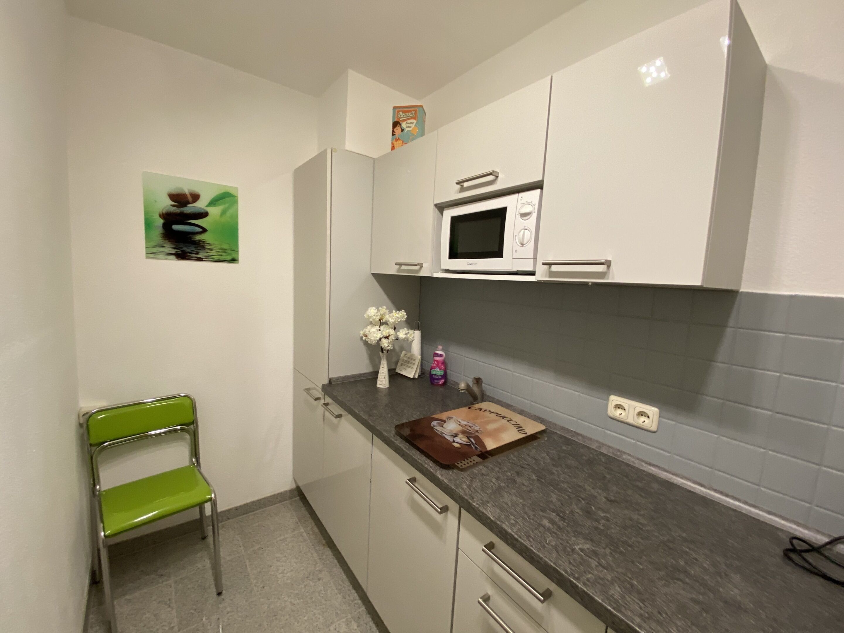 A compact kitchen with white cabinets, a microwave, a green chair, and a small vase with flowers on the counter.