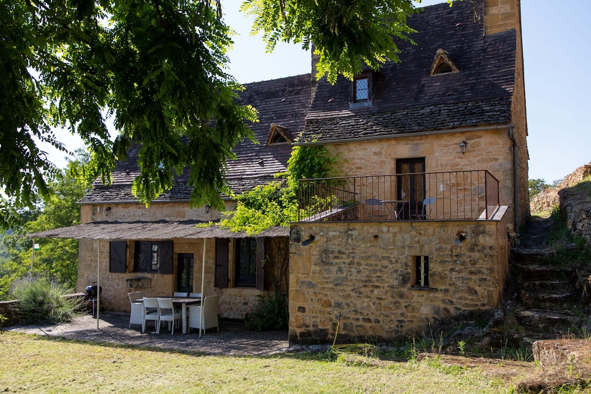 A stone building with a balcony, a table and chairs outside, and a small window.